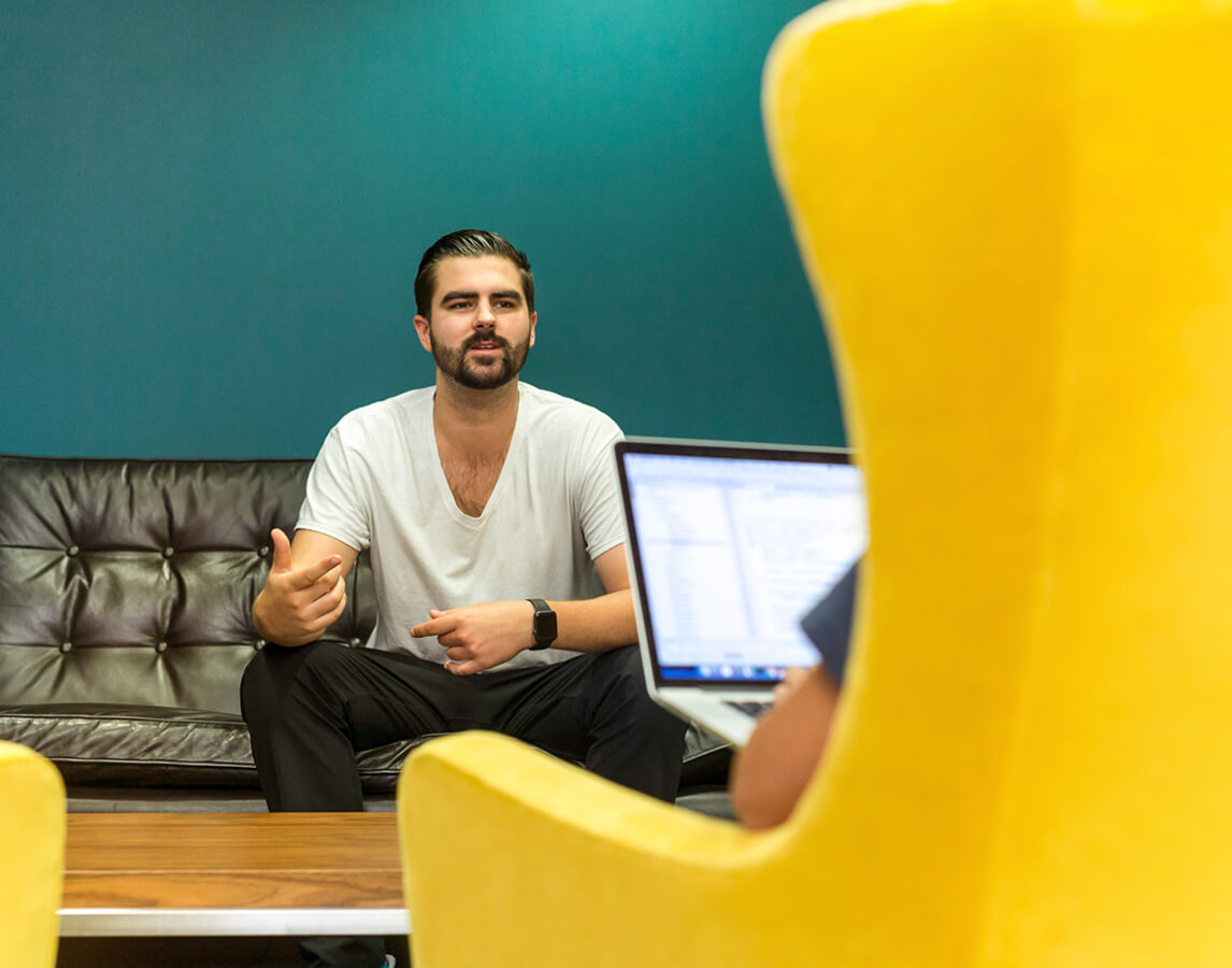 A comfortably dressed man sitting on a couch discussing a project to someone sitting across from him with a laptop.